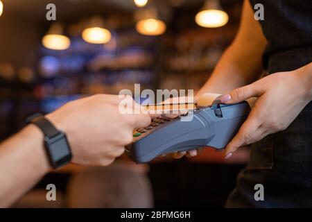 Contemporary young client holding plastic card over payment terminal in waitress hands while paying for drink or lunch in restaurant Stock Photo