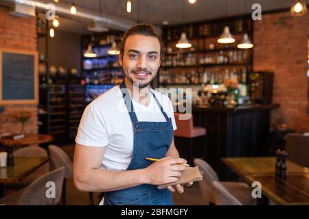 Happy young waiter in white t-shirt and dark blue apron looking at you with toothy smile and writing down your order in restaurant Stock Photo