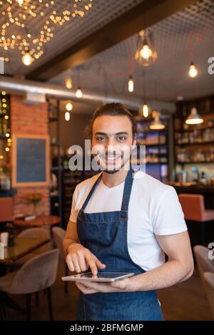 Young cheerful and friendly waiter of classy restaurant scrolling through online orders in digital tablet while waiting for new guests Stock Photo