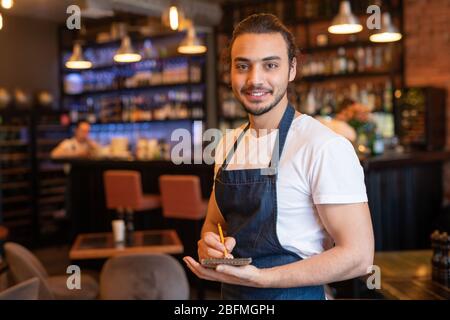 Young smiling waiter in workwear making notes in notepad while standing in front of camera on background of bar counter and interior Stock Photo
