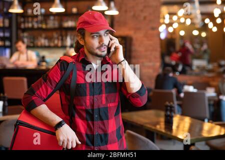 Busy courier in workwear talking to one of clients on mobile phone while asking for address and warning about delivery of ordered food Stock Photo