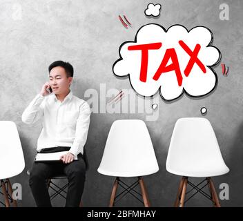 Tax concept. Young Asian businessman sitting on a chair in grey hall Stock Photo