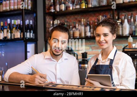Happy young waiter making notes in notepad while his pretty colleague with touchpad standing near by and both looking at you Stock Photo
