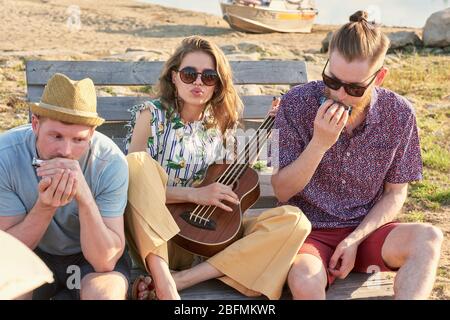 Group of stylish young people resting on wooden bench at the beach and playing musical instruments on warm summer evening Stock Photo