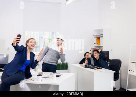Employees having fun in the office, posing, making funny faces and taking selfies with a smartphone. Stock Photo