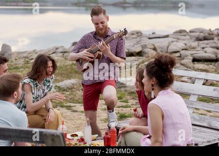 Young bearded man playing the guitar and smiling joyfully while hanging out with friends near lake, sitting around table, eating and talking Stock Photo