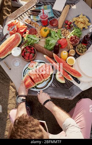 High angle view of unrecognizable man cutting ripe watermelon outdoors while sitting by table with appetizing fresh fruits and vegetables Stock Photo