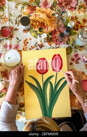 Top view of old woman painting at home in a table full of materials to paint. Art and leisure in the elderly. Painting a picture at home. Stock Photo