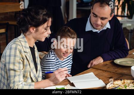A girl doing her homework at the kitchen table with her parents being around. Stock Photo