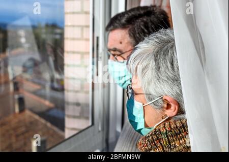 senior couple, with protective face masks, at home looking through the window. Concept of coronavirus quarantine stay home and social distancing. Lockdown people. elderly and retired lifestyle . Stock Photo