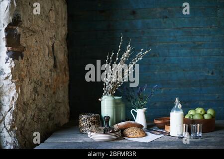 Still life of table served for breakfast with homemade bread, fresh milk in jug and apples in wooden bowl, stack of plates, glasses and napkins Stock Photo