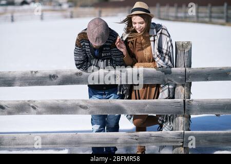 Happy young affectionate couple standing by wooden fence in rural environment while having talk on winter day outdoors Stock Photo