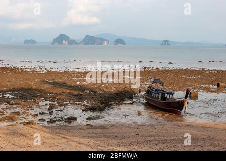 Thai fishing boat stands on the sand at low tide on a rocky shore. A lot of stones in the sand. Cliffs in the sea on the horizon. Horizontal. Stock Photo