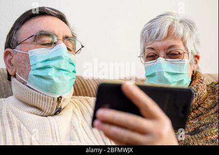 Coronavirus. Stay at home, lifestyle.Cheerful Elderly couple sitting on a sofa on a quarantine at home, making a video call with the smartphone. Senior Couple wearing protective masks . Stock Photo