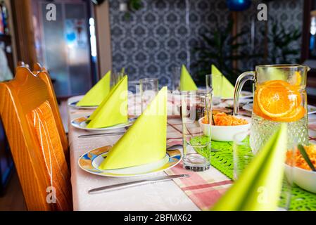 Green napkins arranged on plates in the shape of a triangle, lemonade made of water and orange slices in a large jug, salads visible. Stock Photo
