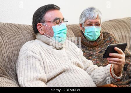 Coronavirus. Stay at home, lifestyle.Cheerful Elderly couple sitting on a sofa on a quarantine at home, making a video call with the smartphone. Senior Couple wearing protective masks . Stock Photo