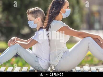 Young woman and a boy sit on a city bench back to back, dressed in light clothes, with one-time face masks on their faces. Stock Photo