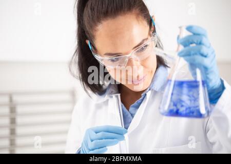 Female chemist wearing protective goggles smiles when holding a test-tube, mixing blue liquid inside the conical flask. Coronavirus COVID-19 concept. Stock Photo