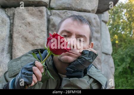 a sad guy in a jacket and leather gloves sitting sniffing a red rose Bud in his hand Stock Photo