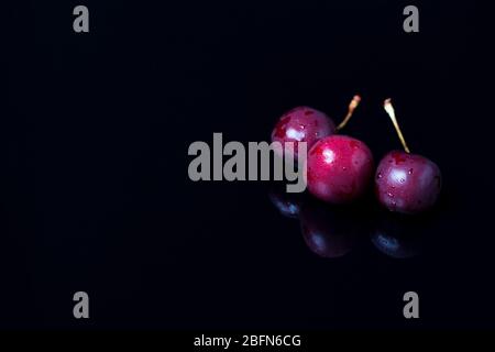 Three cherries on a black mirror background. Reflection, close-up, copy space. Berries, snacks, healthy food. Vegan, vegetarian, diabetic, fruit. Stock Photo