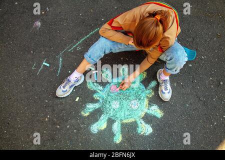 Teenage girl sits on a skateboard and draws coronavirus with chalk on the pavement Stock Photo