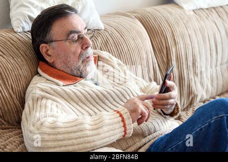 Elderly Man Using Cellphone Sitting On Sofa At Home, during Coronavirus quarantine  Stock Photo