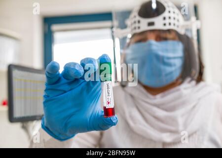 COVID-19 outbreak. Laboratory specialist in protective face mask and shield. SARS-CoV-2 positive result test tube in hand in blue glove. Health care. Stock Photo