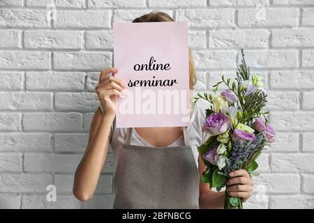 Woman holding paper with text ONLINE SEMINAR and bouquet on brick wall background. Florist and floral design tutorial concept. Stock Photo