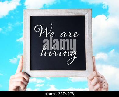 Labor market concept. Female hands holding chalkboard with text WE ARE HIRING on sky background Stock Photo
