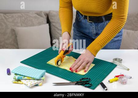 woman in a yellow cuts the fabric. there are quilting tools on the table. patchwork knife, scissors, lined cutting mat, self-locking, thread, centimet Stock Photo