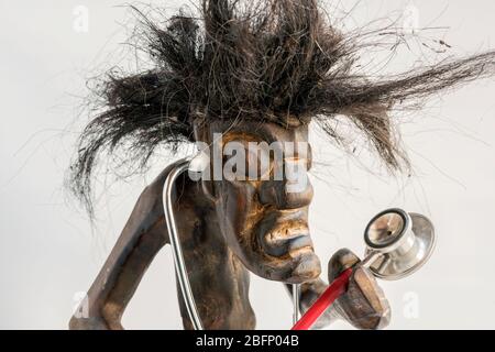 Wooden carved figure of primitive man wearing straw skirt & long black tousled hair, with stethoscope. Concept; Aspiration, Ambition, Social mobility. Stock Photo