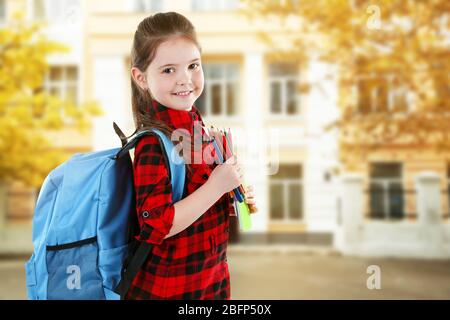 Little girl with blue back pack holding pencils on blurred school building background Stock Photo