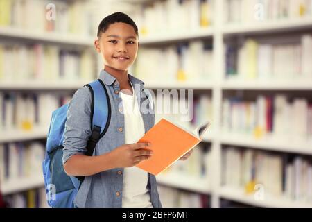 Cute boy with backpack holding book on blurred book shelves background. Library concept. Stock Photo