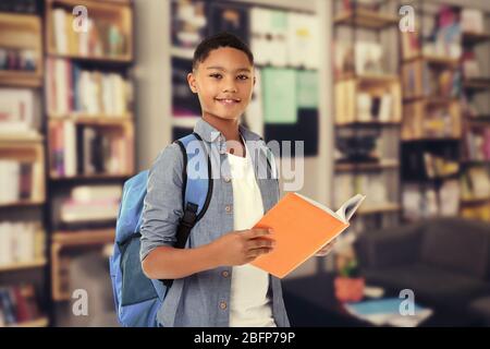 Cute boy with backpack holding book on blurred book shelves background. Library concept. Stock Photo