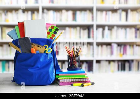 Blue backpack with school supplies on wooden table against blurred book shelves. School concept. Stock Photo