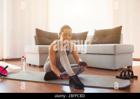 Young woman doing sport workout in room during quarantine. Sit on mat and tuing shoes laces. Preparing for home workout. Stock Photo