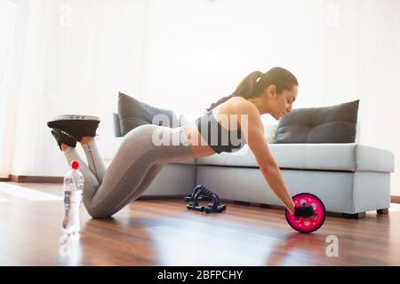 Young woman doing sport workout in room during quarantine. Side view of woman using abdominal exercise roller for stretching forward. Stand on knees Stock Photo
