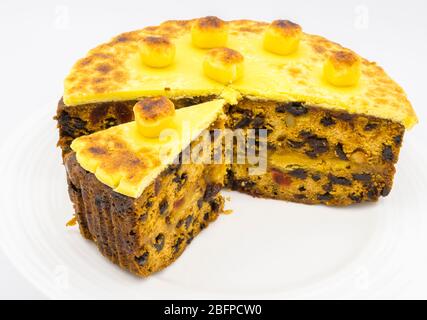 Traditional home baked simnel cake with a cut slice on a white plate for Easter - fruit cake topped with toasted yellow marzipan topping and balls Stock Photo