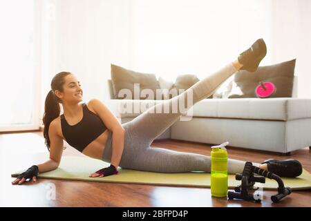 Young woman doing sport workout in room during quarantine. Girl lying on hip side and hold left leg up. Stretching low body part. Exercising alone in Stock Photo