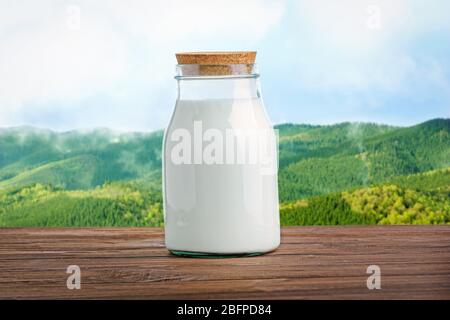 Glass bottle of milk on wooden table against blurred landscape background. Dairy concept. Stock Photo