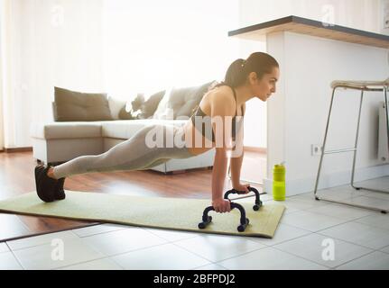 Young woman doing sport workout in room during quarantine. Picture of strong fitness model stand in plank position using push up stands hand bar Stock Photo