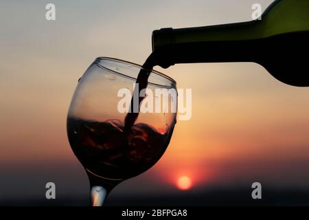 Red wine pouring from a bottle into the glass on beautiful sunset background, orange sun is shining through the clouds. Concept of celebration Stock Photo