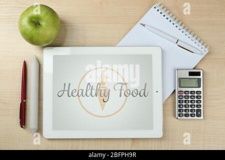 Tablet, apple and school stationery on wooden background. Text HEALTHY FOOD on screen Stock Photo