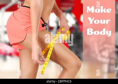 Weight loss motivation concept. Young woman measuring thigh with tape in gym, closeup. Text MAKE YOUR BODY on background Stock Photo