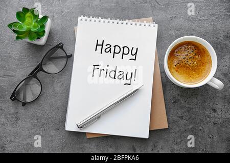 Notebook with text HAPPY FRIDAY, glasses and cup of coffee on gray background Stock Photo