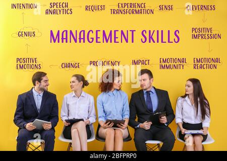 Young people with clipboards sitting on chairs and diagram of MANAGEMENT SKILLS against yellow wall background Stock Photo