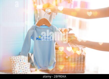 Woman holding baby bodysuit with text MY FIRST HANUKKAH at home Stock Photo