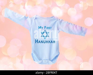 Baby bodysuit with text MY FIRST HANUKKAH on blurred lights background Stock Photo