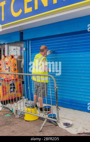 A painter and decorator painting the metal shutter on the exterior of a shop wearing a high visibility waistcoat Stock Photo