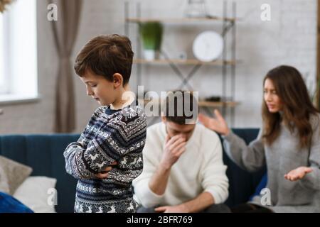 Sad little boy feels upset while his parents fighting at background. Mother and father quarrel. Family conflicts and divorce lead to children's Stock Photo
